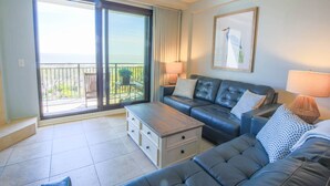 Ein modernes Wohnzimmer mit einem großen Schiebeglasfenster, das auf einen Balkon führt, einem schwarzen Ledersofa, einem Holztisch und einem Bild an der Wand.