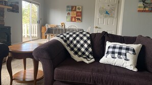 Ein gemütliches Wohnzimmer mit einem dunkelbraunen Sofa, einer karierten Decke und einem Kissen mit Bärchendruck. Im Hintergrund sind ein Holztisch und Stühle zu sehen, und an der Wand hängt eine Karte.