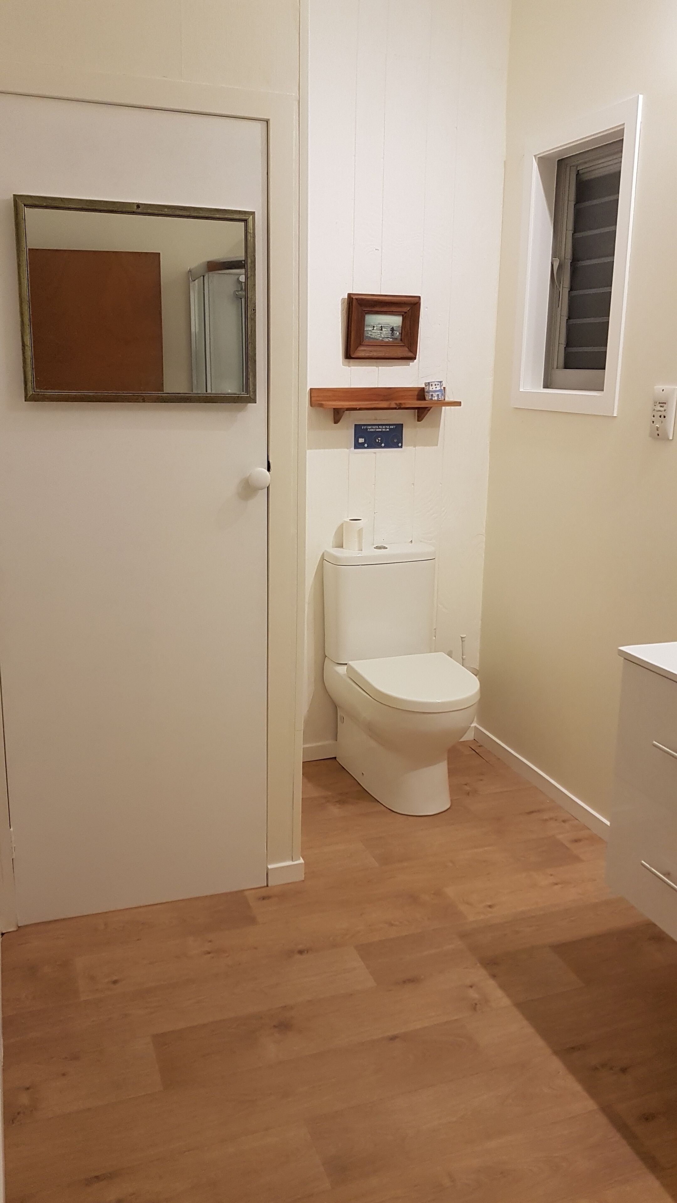 A bathroom with a toilet, a mirror, a shelf with a picture frame, and a window.