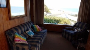 Ein gemütliches Zimmer mit einem gestreiften Sofa und Sesseln, großen Fenstern mit Blick auf Strand und Meer, und Holzvertäfelung.