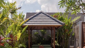 A pool with a wooden gazebo and a bench, surrounded by tropical plants.