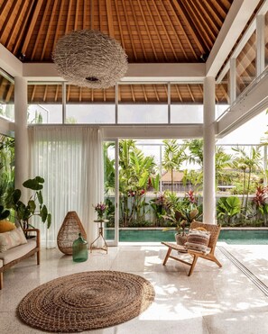 Un vaste espace intérieur doté d’un plafond en bois, de grandes fenêtres et offrant une vue sur une piscine et un espace verdoyant à l’extérieur.