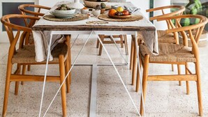 A dining area with a table set for four, surrounded by wicker chairs, a potted plant, and a variety of fruits and nuts on the table.
