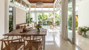 A spacious room with a wooden dining table, wicker chairs, and a bench with cushions. There is a large window providing a view of lush greenery outside.
