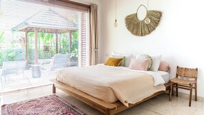 A bedroom with a large bed, a chair, a hanging light fixture, and a view of greenery outside.