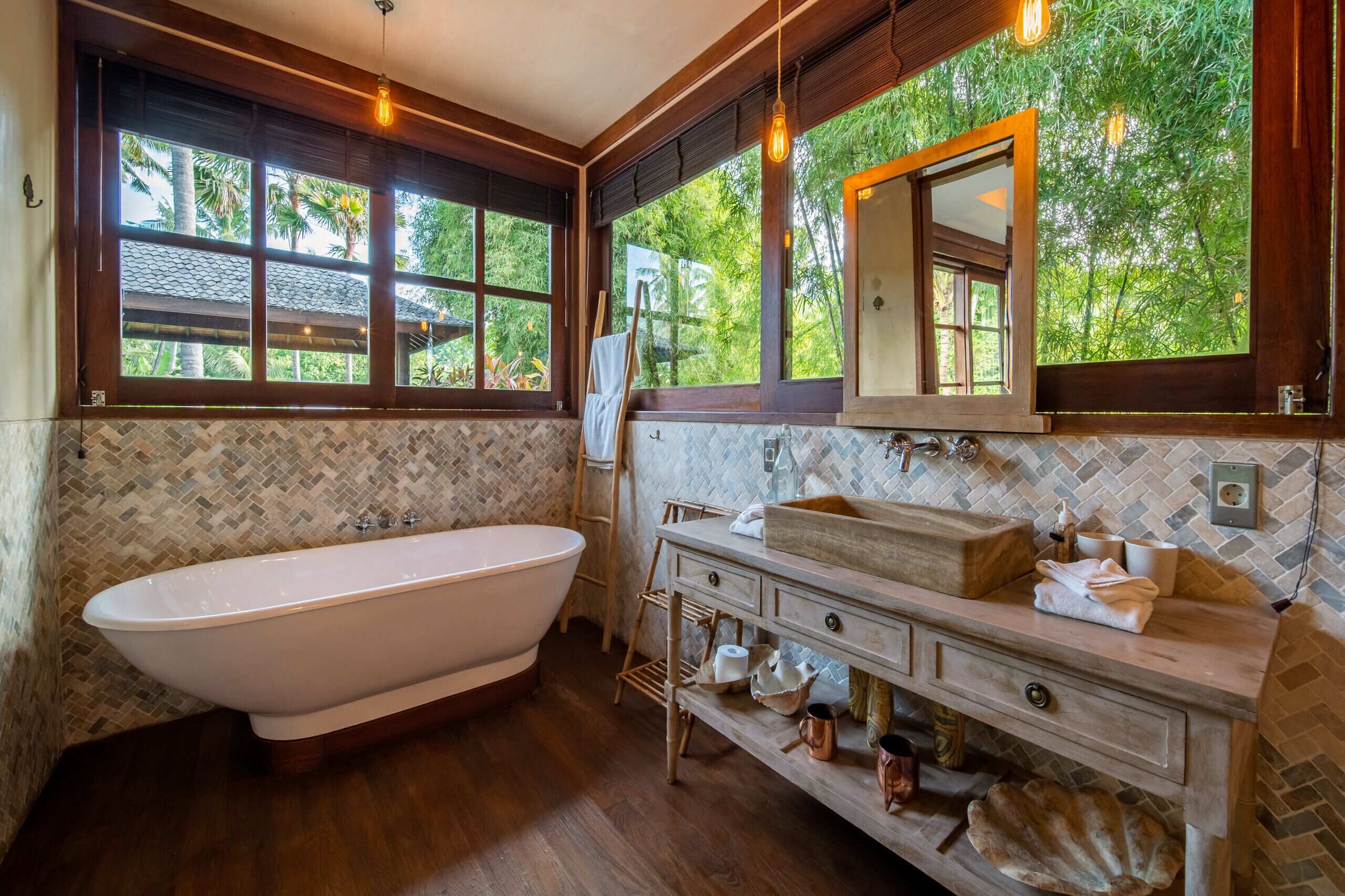 Ein Badezimmer mit freistehender Badewanne, einem Holzbadezimmer-Möbelstück mit Spiegel und Blick ins Grüne durch große Fenster.