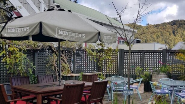 An outdoor patio area with wooden tables and chairs, covered by umbrellas, and surrounded by potted plants. - Hampden Hotel (Murchison)