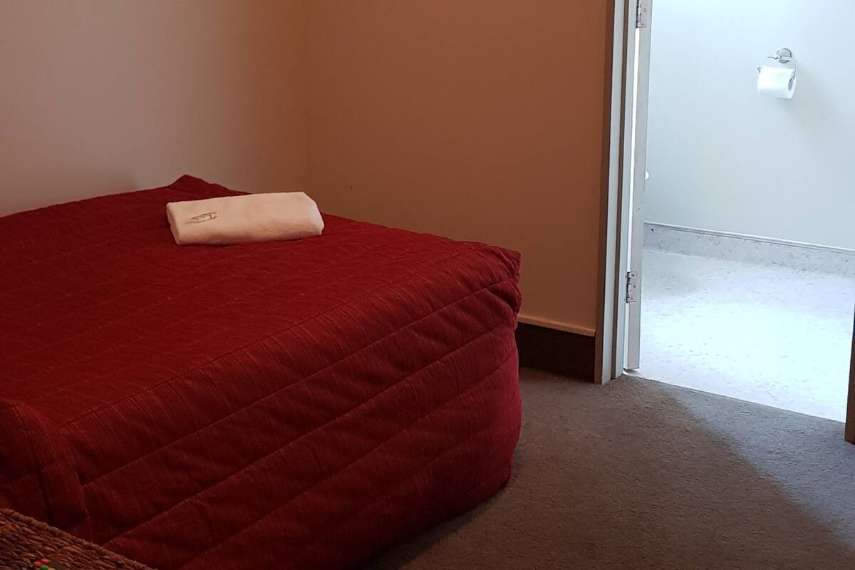 A room with a red bed, a white towel, and a remote control on a small table.