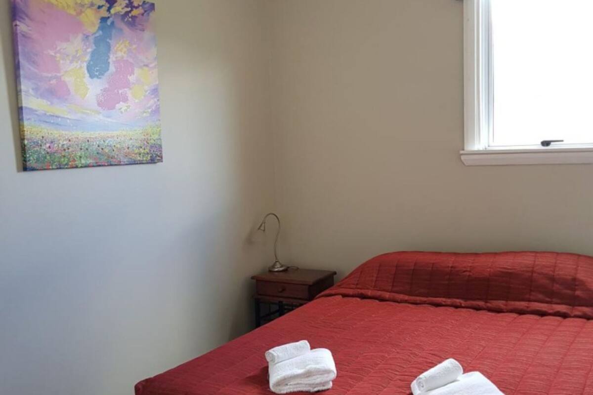 A room with a red bedspread, two neatly folded white towels on the bed, a small wooden nightstand with a lamp, and a colorful abstract painting on the wall.