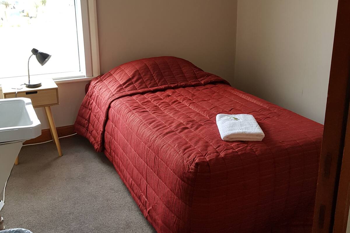 A small, neatly made bed with a red quilt, a white bedside table with a lamp, and a window with a view of the outside.