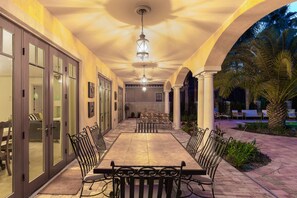 Un espacio al aire libre cubierto con una mesa larga y sillas, rodeado de puertas de cristal y palmeras.