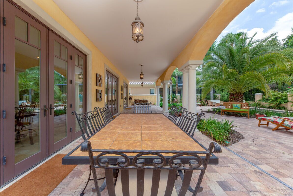 A spacious outdoor dining area with a long table and chairs, surrounded by glass doors and a covered walkway.