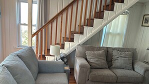 Una sala de estar con un sofá seccional gris, un sofá gris más pequeño, una escalera de madera y un ventilador en el suelo.
