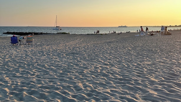 Una playa con arena, un velero a lo lejos y algunas personas caminando.