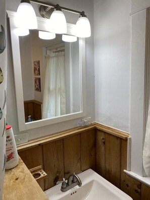 Un baño con lavamanos blanco, paneles de madera, un espejo grande con tres luces y cortina de ducha.