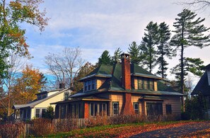 Una casa de dos pisos con techo verde y chimenea de ladrillo, rodeada de árboles y una cerca de madera.