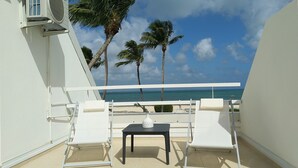 A balcony with two white lounge chairs and a small table, overlooking a beach with palm trees and the ocean. - Friendly Beach House (4160 nettle baie beach club)