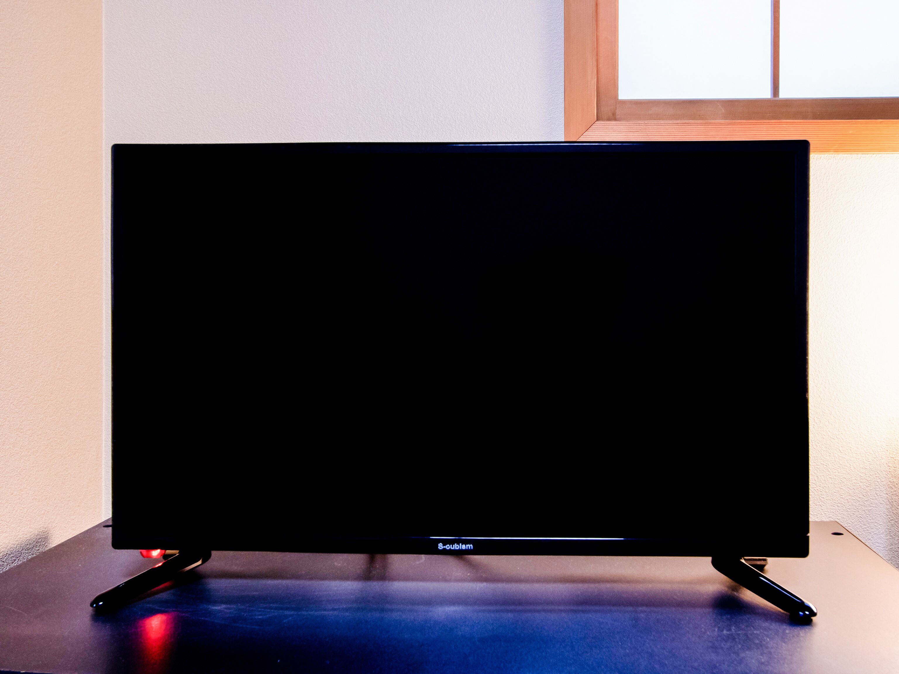 Un téléviseur à écran plat, posé sur un meuble avec un cadre en bois, est placé sur une table adossée à un mur près d’une fenêtre.