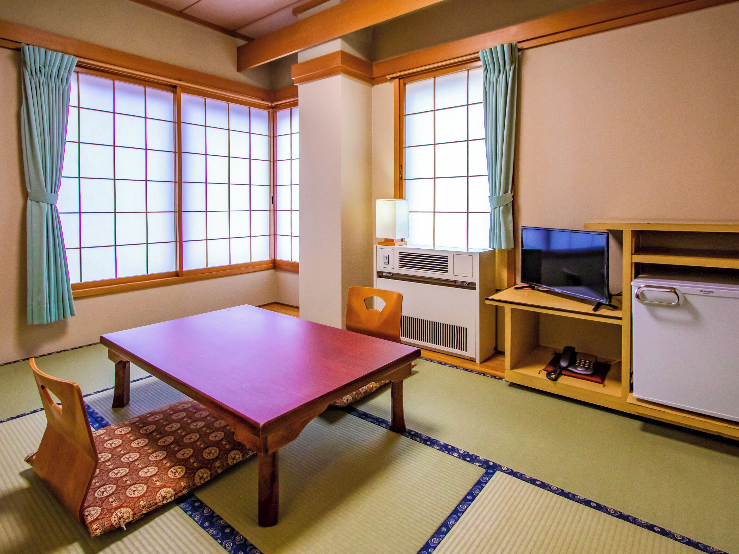 A traditional Japanese room with tatami flooring, a wooden table, a television, and a refrigerator.