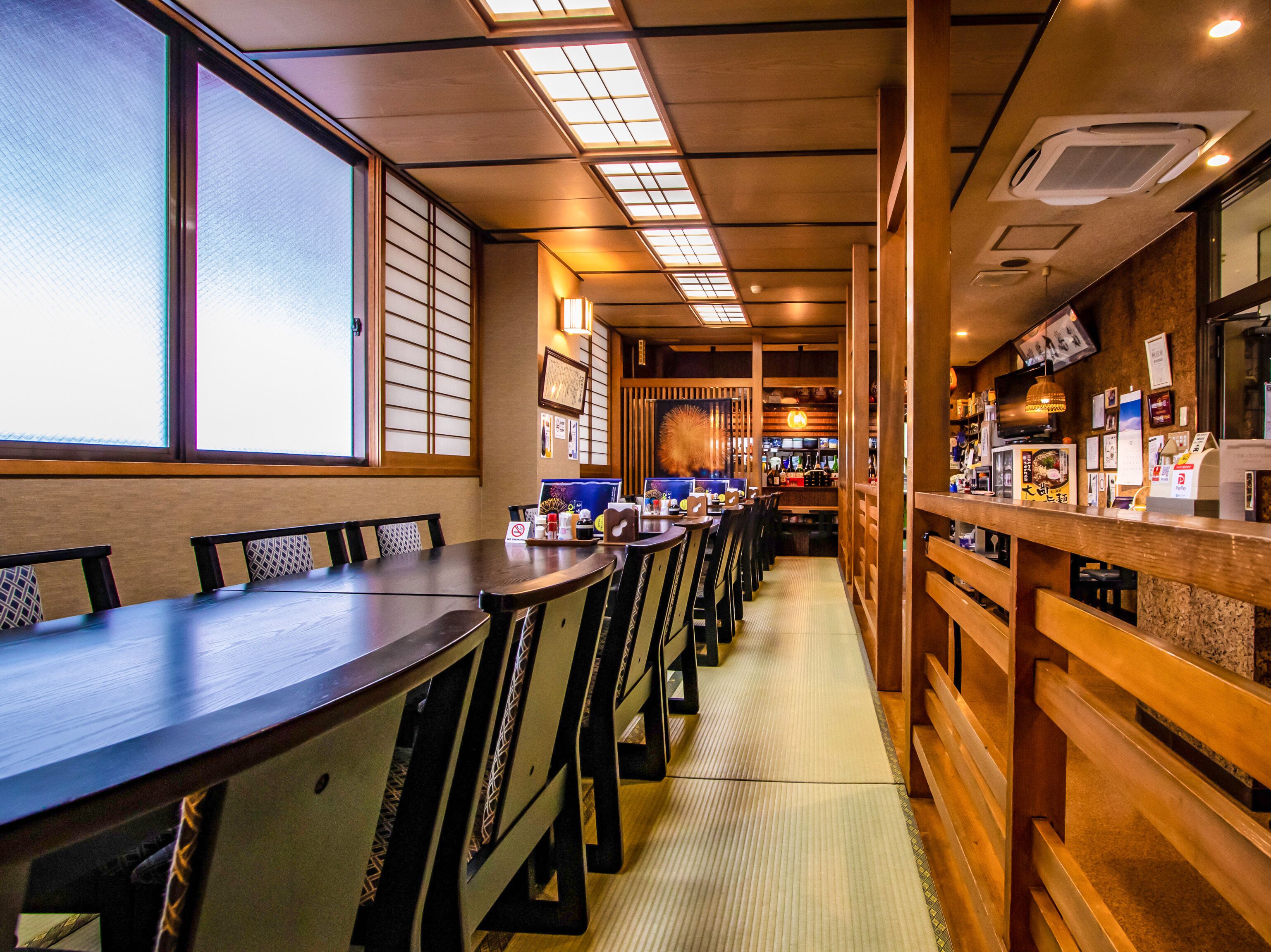 A restaurant with wooden tables and chairs, a counter with a menu, and a view of the street through large windows.
