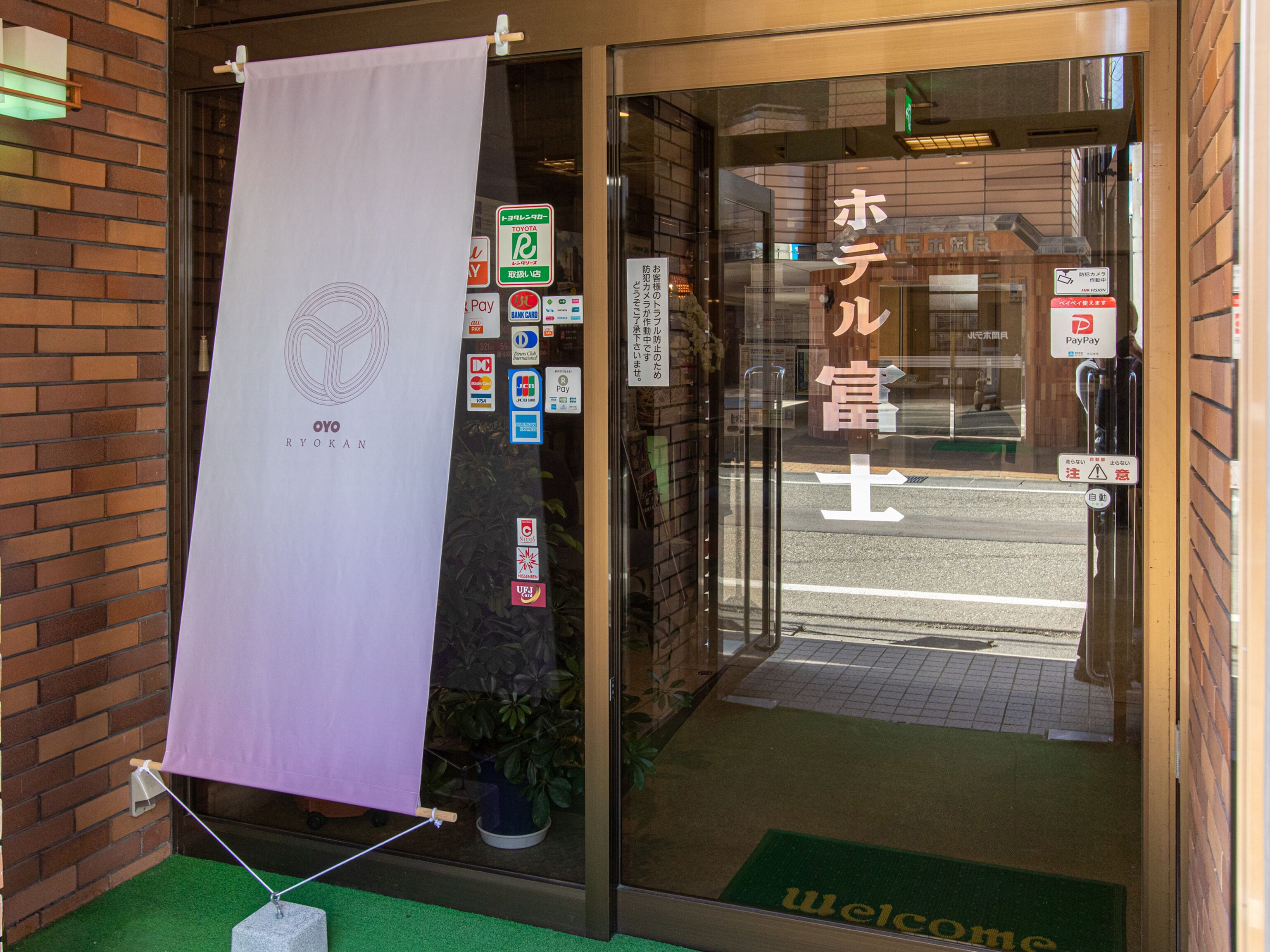 A hotel entrance with a 'Welcome' mat, a 'Pay' sign, and a 'TOYOTA' parking sign.