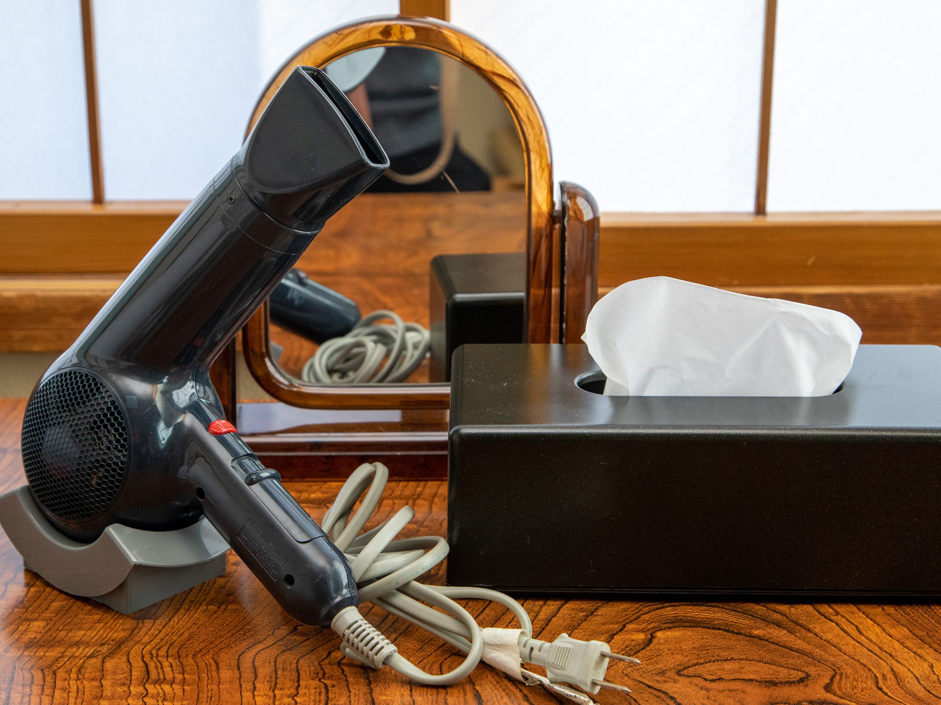 A hair dryer on a wooden surface with a mirror, a tissue box, and a power cord.