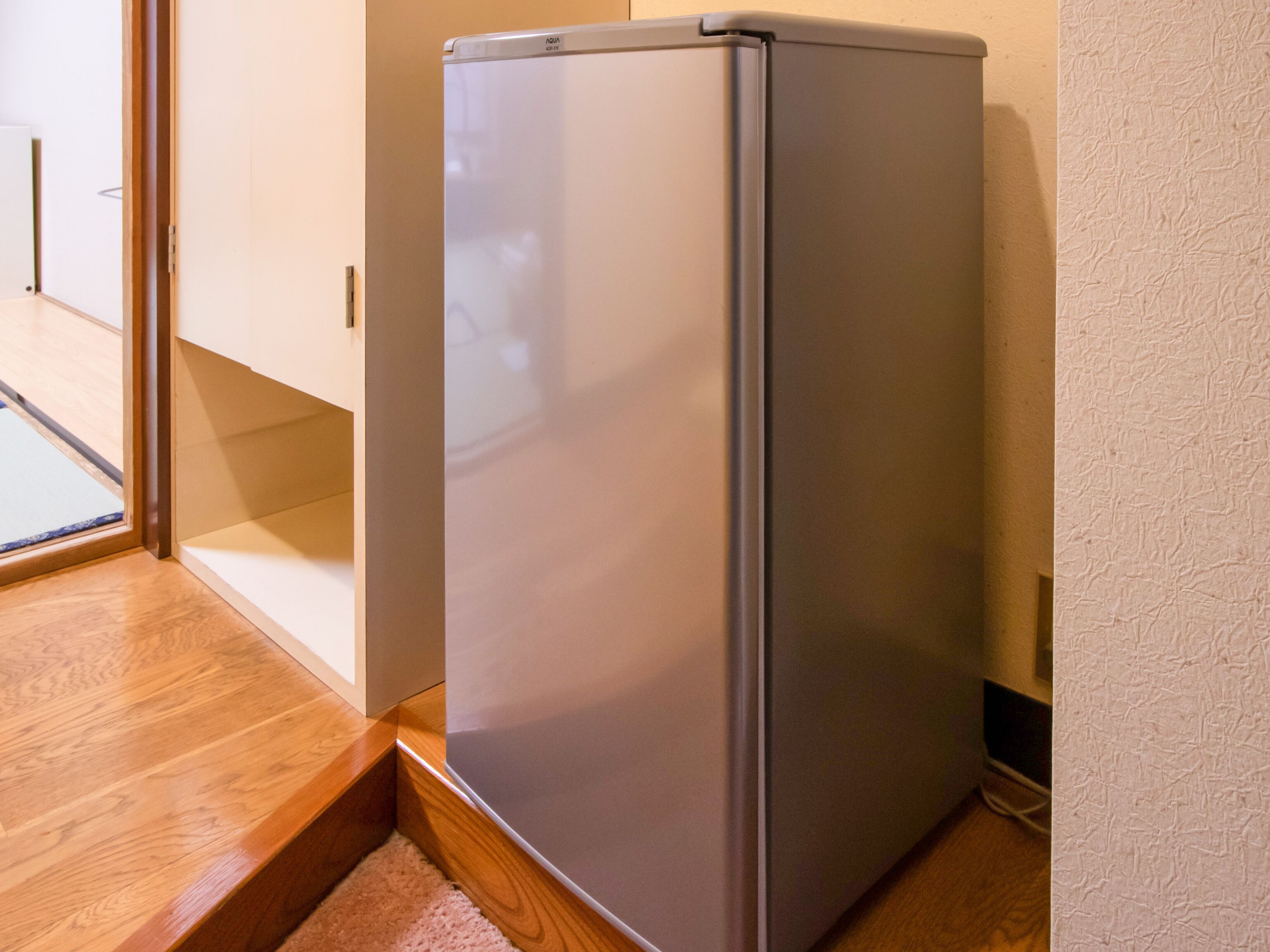 A compact, modern refrigerator in a corner of a room with wooden flooring and a glass door leading to another area.