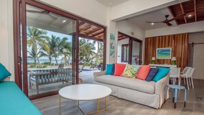A spacious living area with a large sliding glass door leading to a balcony, a beige sofa with colorful cushions, a round white table, and a small blue stool. - PRIVATE BEACHFRONT PARADISE with COOK & POOL = Luxury on the Sea! (San Pedro)