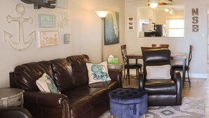 Una sala de estar con un sofá de cuero, reposapiés y decoración en la pared. Se ve un comedor con mesa y sillas al fondo.