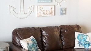 Un sofá de cuero marrón con almohadones decorativos, un reposapiés azul y arte de pared que incluye un ancla y citas motivacionales.