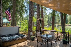 A hot tub on a deck with a table and chairs, surrounded by tall trees and a view of a lake.