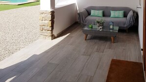 Una veranda coperta con un muro in pietra, una tenda bianca, un divano grigio con cuscini verdi, un tavolino da caffè in vetro e una piscina sullo sfondo.