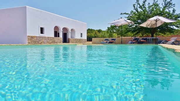 Una piscina con acqua cristallina, un edificio bianco con dettagli in pietra e lettini prendisole con ombrelloni.