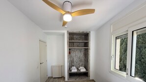 A room with a ceiling fan, a wooden floor, a white bed, a white bench, a radiator, a wooden wardrobe, and a window with a view of trees. - Agi Rescator 219 Apartamento for rent in Roses Girona. (Rosas / Roses)