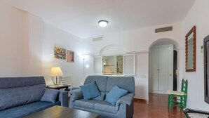 Sala de estar con un sofá gris, una mesa de centro de madera y una lámpara. Hay un sillón verde y una mesita con una lámpara. Las paredes son blancas y el piso está cubierto de baldosas.