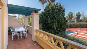 Un patio con una mesa y sillas blancas, un toldo verde y una piscina al fondo.