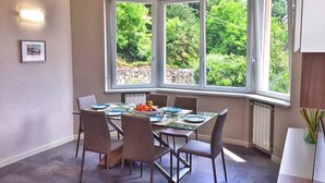 Un coin repas avec une table dressée pour quatre personnes, de grandes fenêtres donnant sur un paysage verdoyant et un radiateur fixé au mur.