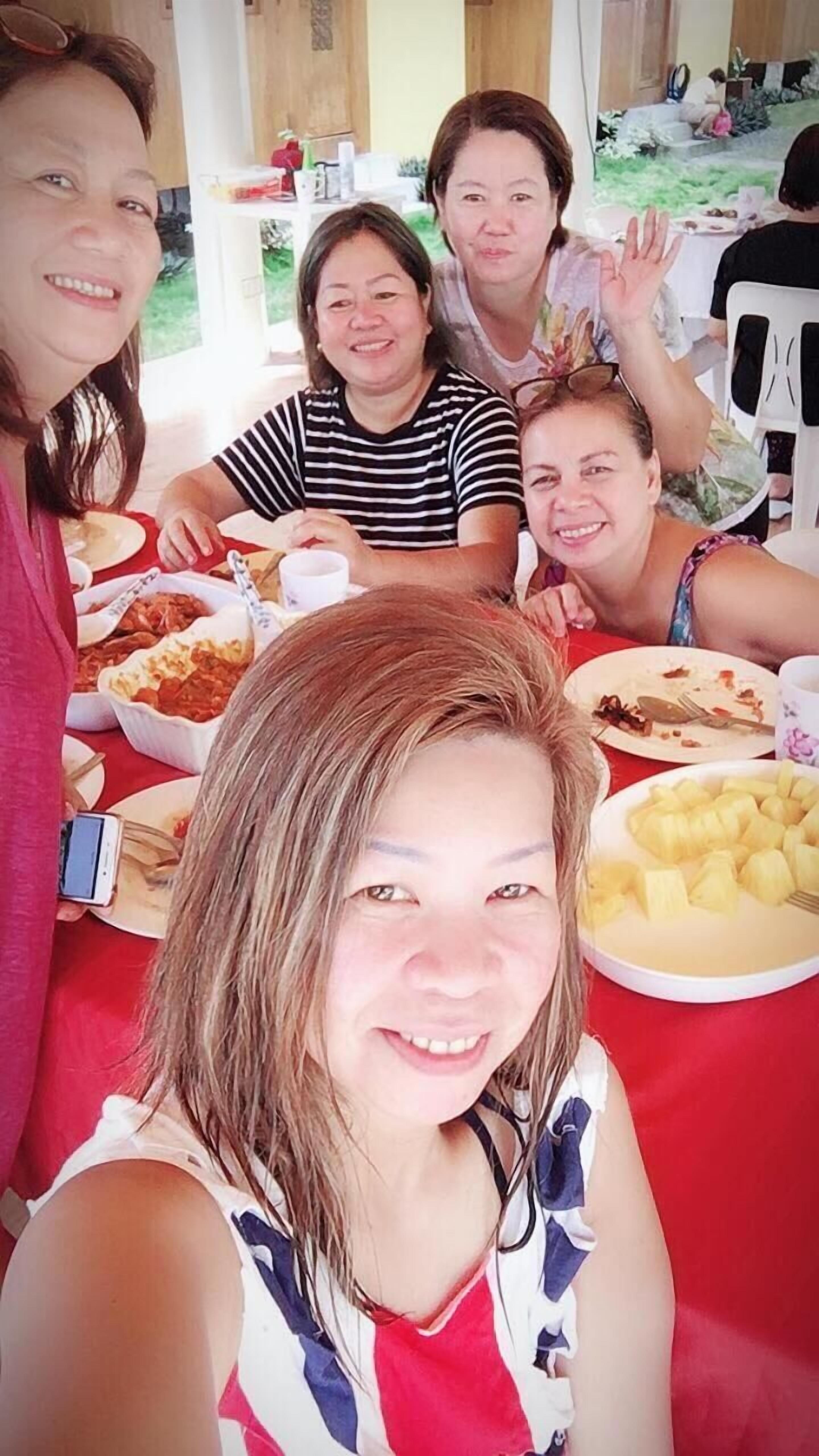 Un grupo de personas sentadas alrededor de una mesa con varios platos, incluyendo un plato de trozos de piña, en una habitación con mantel rojo.