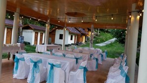 An outdoor event space with tables covered in white tablecloths and blue sashes, under a wooden canopy with string lights. - Aundanao Oasis Beach (Samal)