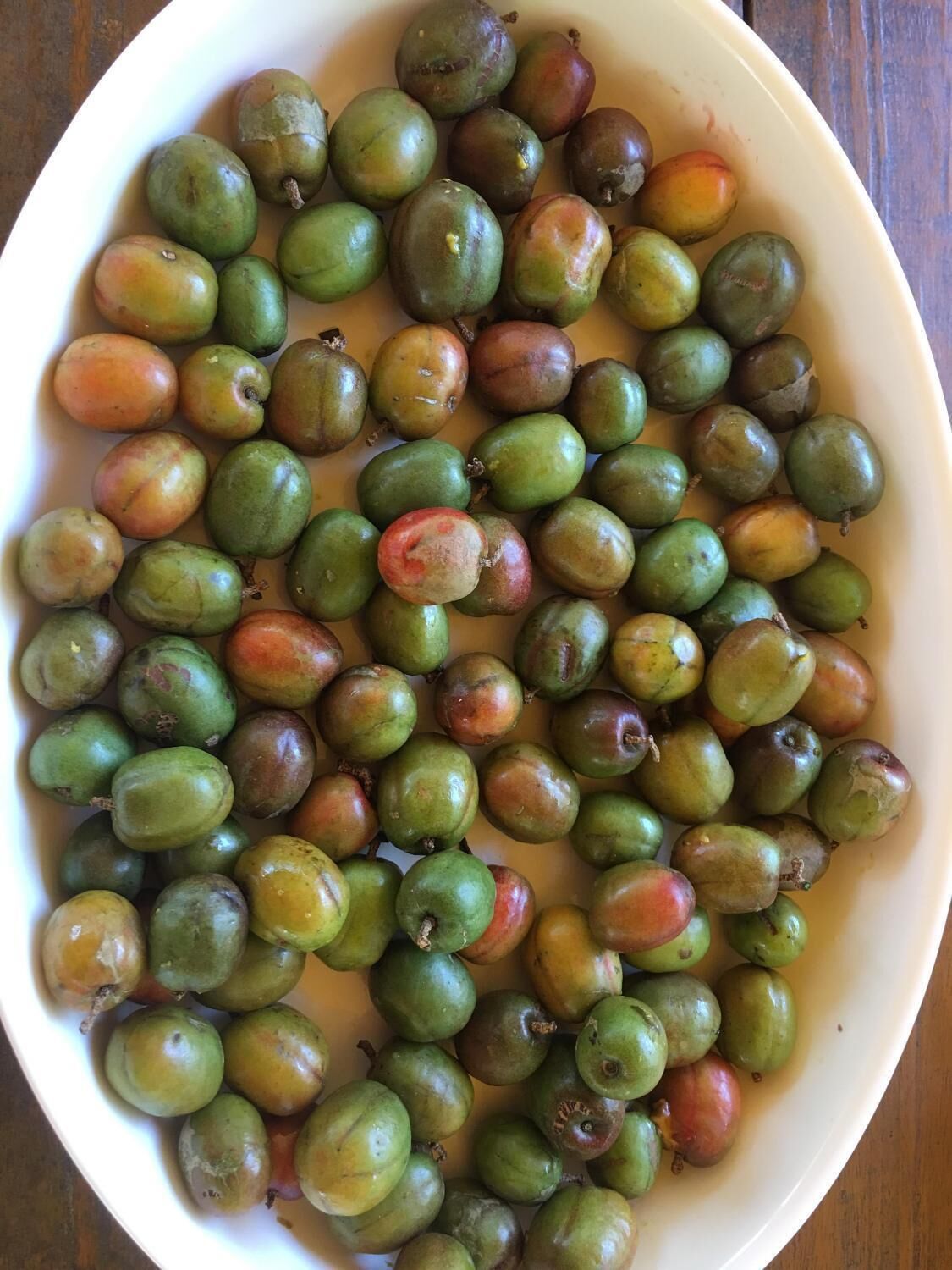 A bowl of green and red fruits, likely guavas, on a wooden surface.