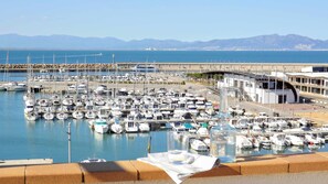 Una marina con numerosos barcos amarrados, un puente al fondo y una vista despejada del mar y las montañas.