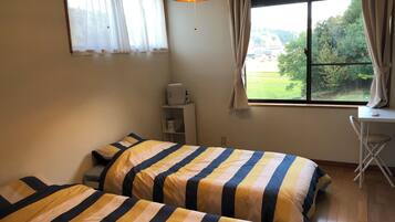 A bedroom with two beds, a desk, a chair, and a window with a view of greenery.