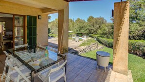Un espace extérieur abrité, aménagé pour les repas, avec une table et des chaises en verre, une petite table ronde, et une vue sur un jardin avec une piscine et des arbres.