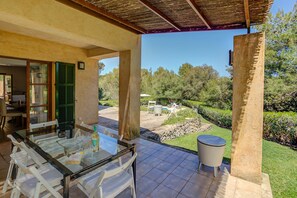 Un espace extérieur abrité, aménagé pour les repas, avec une table et des chaises en verre, une petite table ronde, et une vue sur un jardin avec une piscine et des arbres.