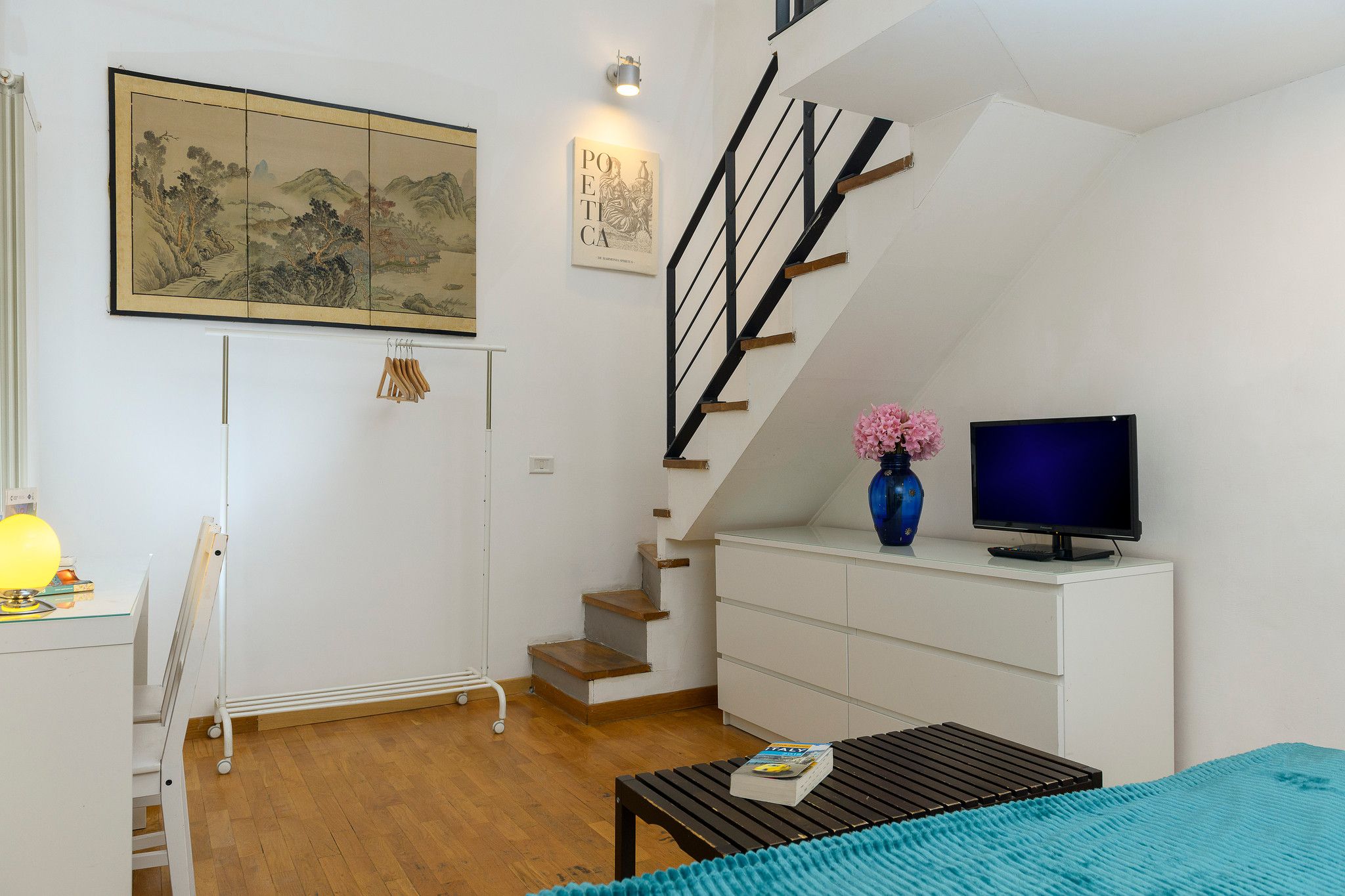 A room with a wooden floor, a white desk, a blue vase with flowers, a television, and a staircase.