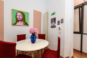 Une pièce avec une table ronde blanche, des chaises rouges, un vase bleu contenant des fleurs roses et divers éléments décoratifs sur les murs, notamment une célèbre peinture et un panneau de sortie vert.