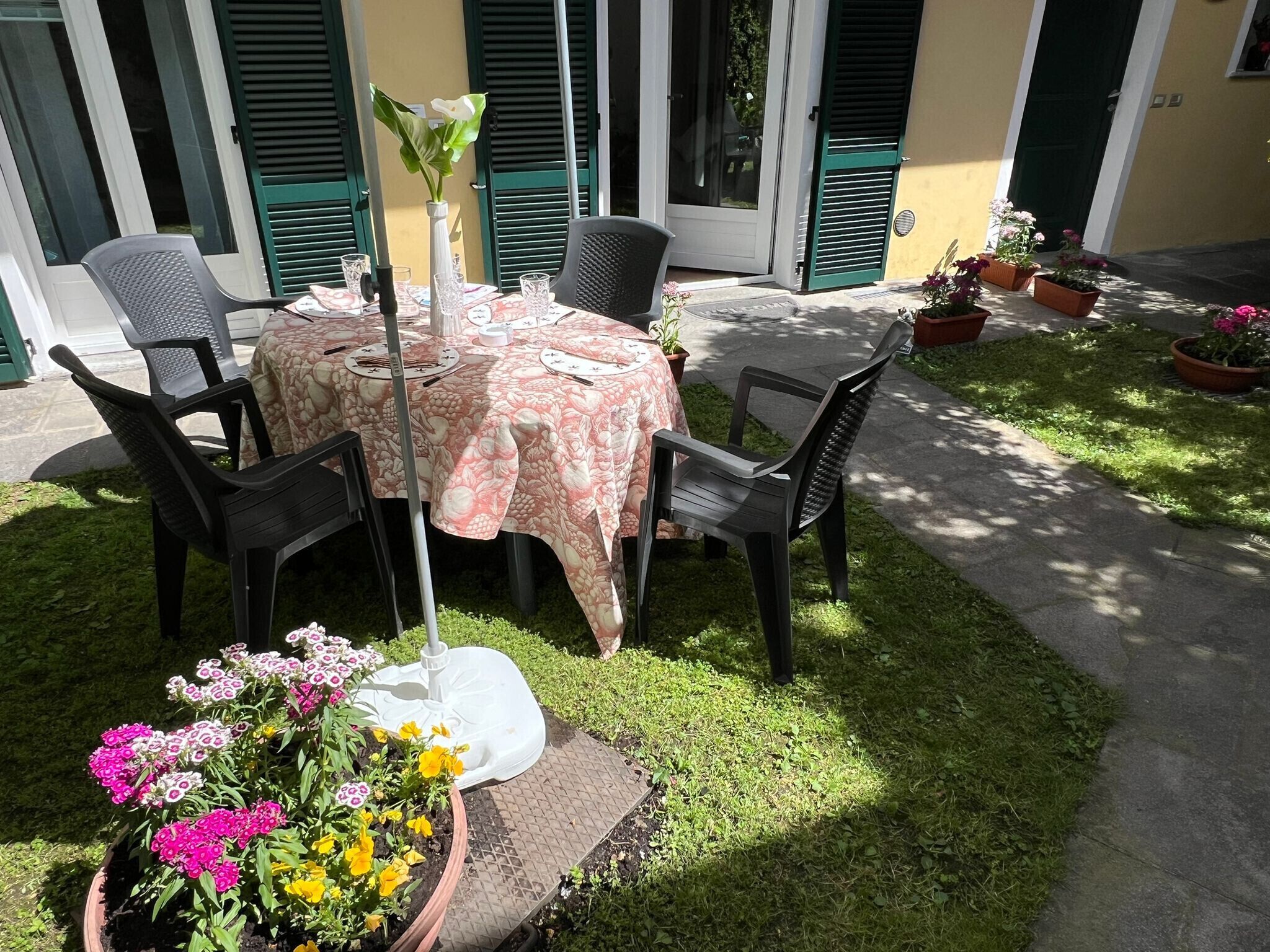 An outdoor dining area with a table set for four, surrounded by chairs. There are potted plants and a decorative fountain in the foreground.