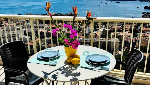 A round table with a yellow vase of flowers and lemons, two black chairs, and a view of the sea and buildings in the background. - Sunset paradise in Villefranche (Villefranche-sur-Mer)