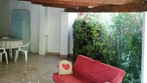 A covered patio area with a red sofa, a small table, and a chair. There is a blue door and a green plant in the background. - The blue holiday home of La Pervinca (Villassunta)