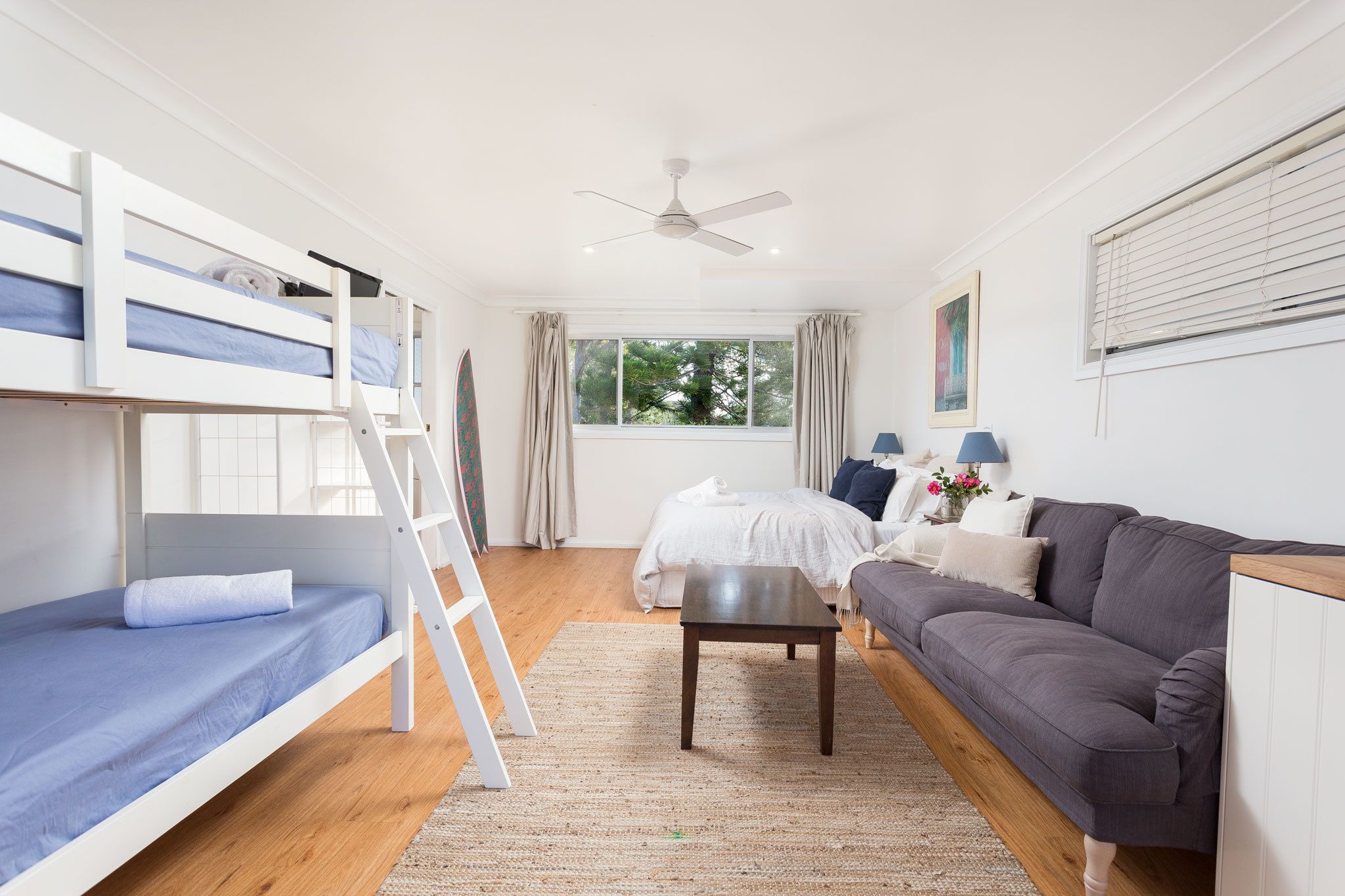 A room with bunk beds, a sofa, a table, a ceiling fan, and a window with curtains.